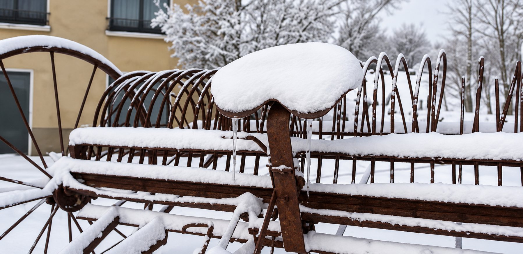 Un ancien râteau agricole à dents métalliques et à grande roue en bois, recouvert d'une épaisse couche de neige devant un bâtiment.