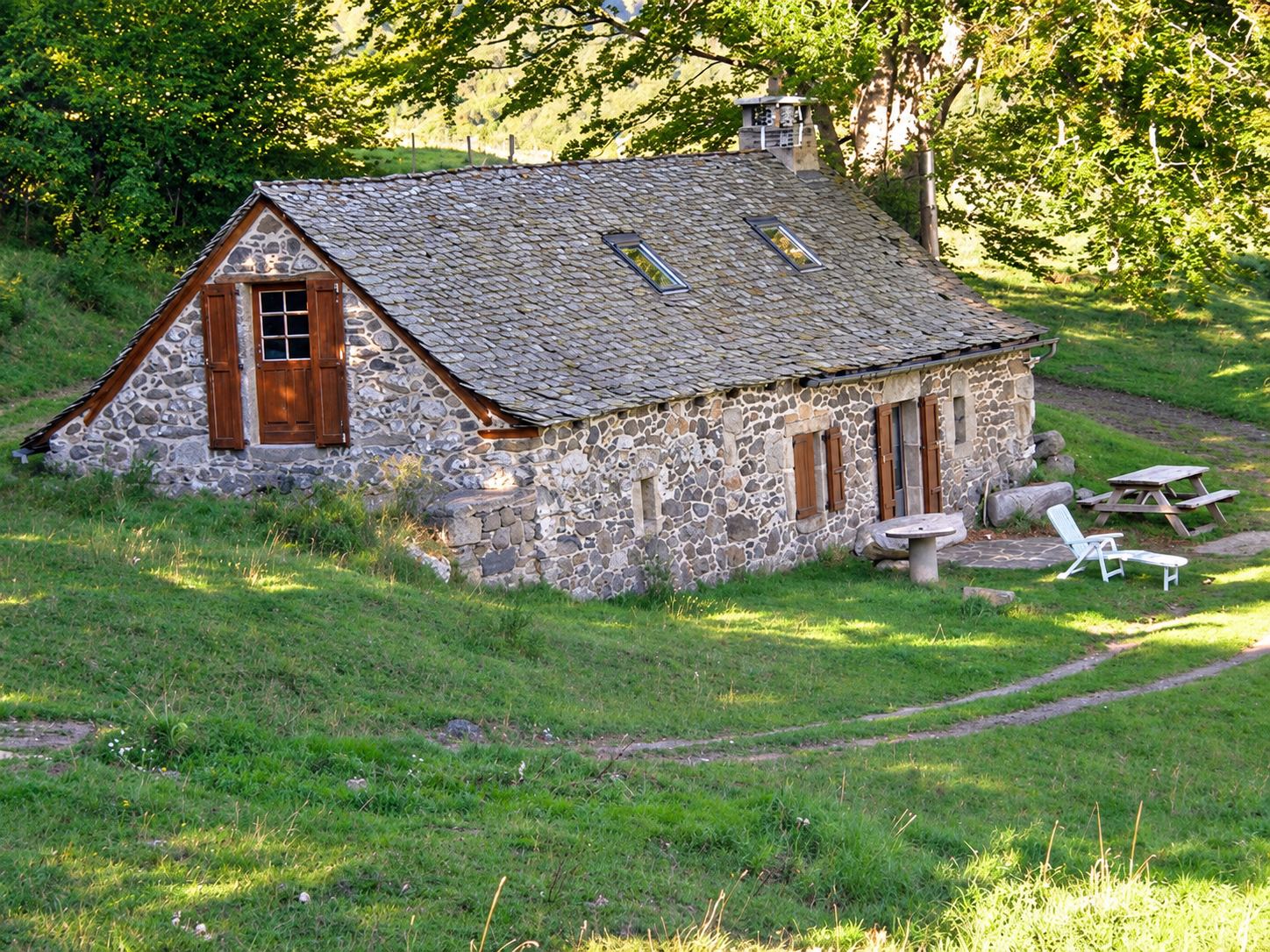 Bâtisse en pierre au toit de lauzes et aux volets bruns, entourée d'herbe, avec une table de pique-nique et une chaise longue blanche.