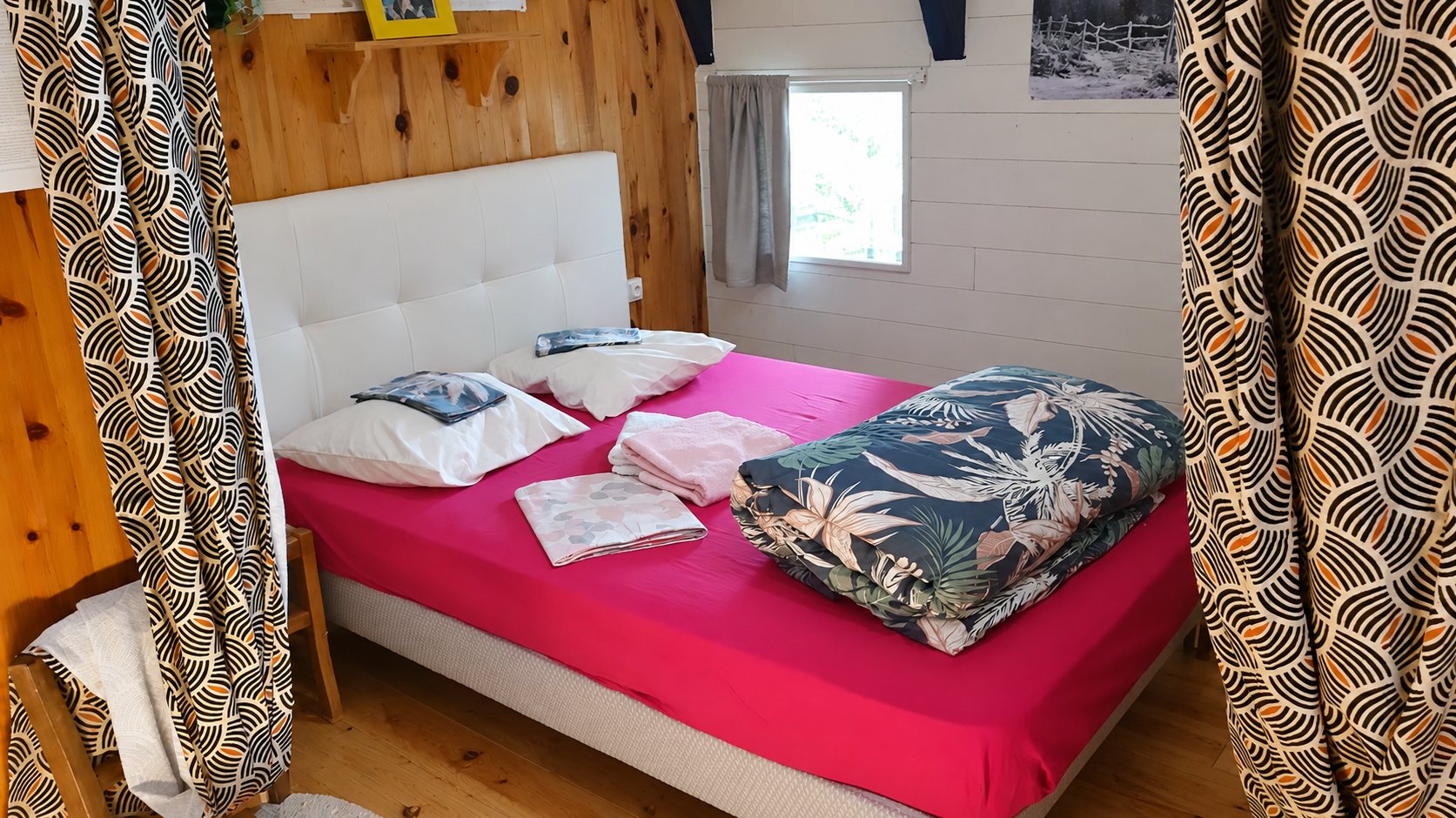 Chambre aux murs en bois avec un lit double aux draps roses, une couette à motif tropical et des rideaux graphiques de chaque côté.