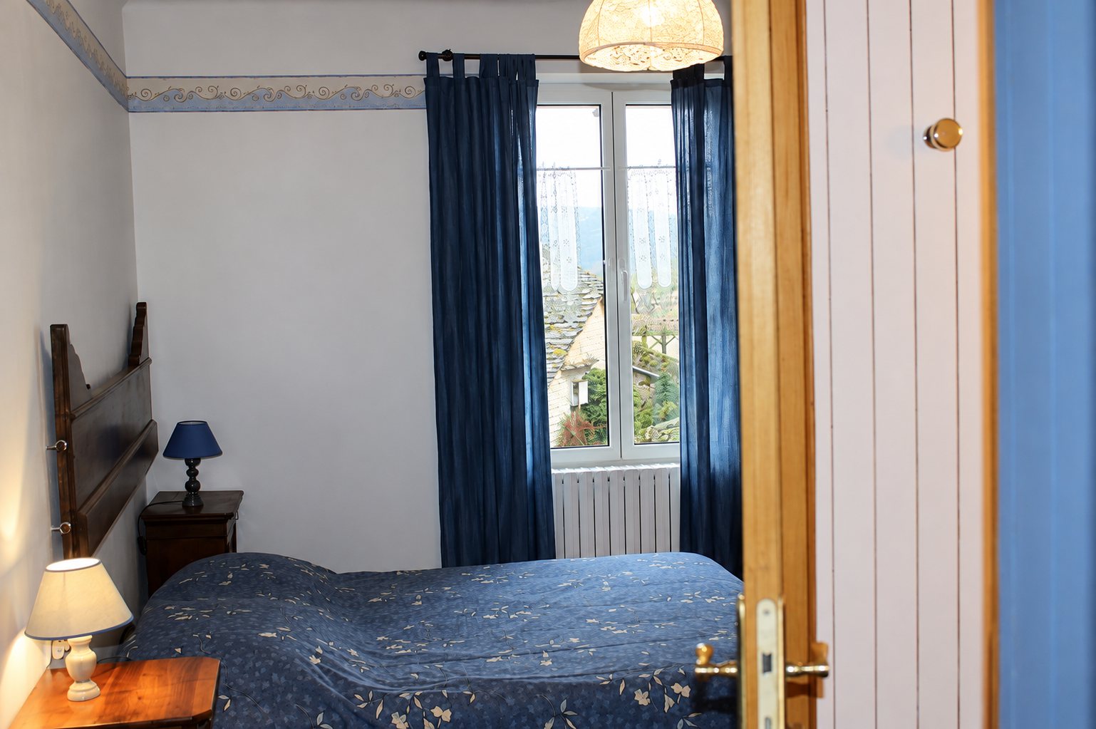 Chambre avec un lit double couvert d'un plaid bleu à motifs, des rideaux bleus, deux lampes de chevet et une fenêtre donnant sur des toits de pierre.