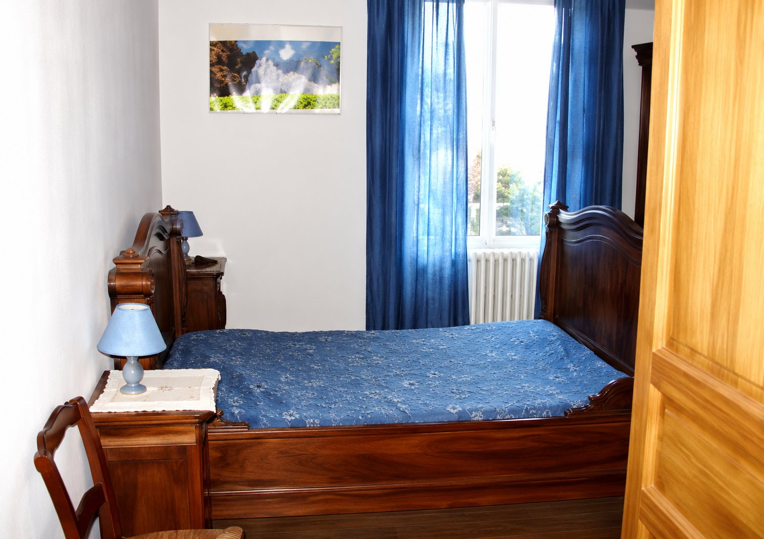 Une chambre aux murs blancs avec un lit en bois recouvert d'un couvre-lit bleu, une lampe de chevet, une commode et un rideau bleu à la fenêtre.