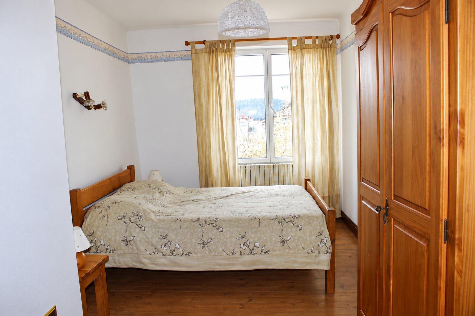 Chambre avec un lit double en bois recouvert d'un couvre-lit à motifs floraux, des rideaux beiges devant la fenêtre et une armoire en bois à droite.
