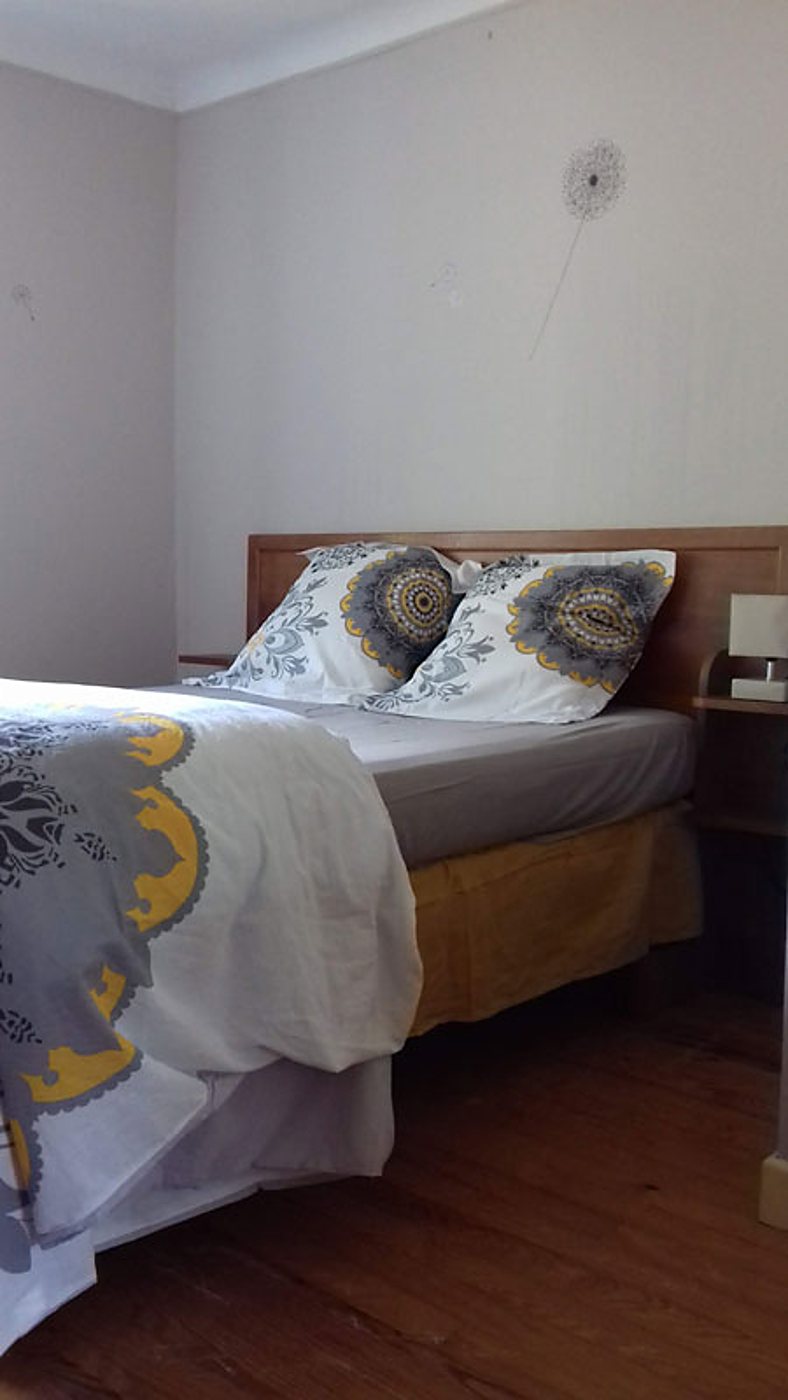 Chambre aux murs clairs avec un lit double habillé de linge blanc à motifs gris et jaunes, une tête de lit en bois et un parquet foncé.