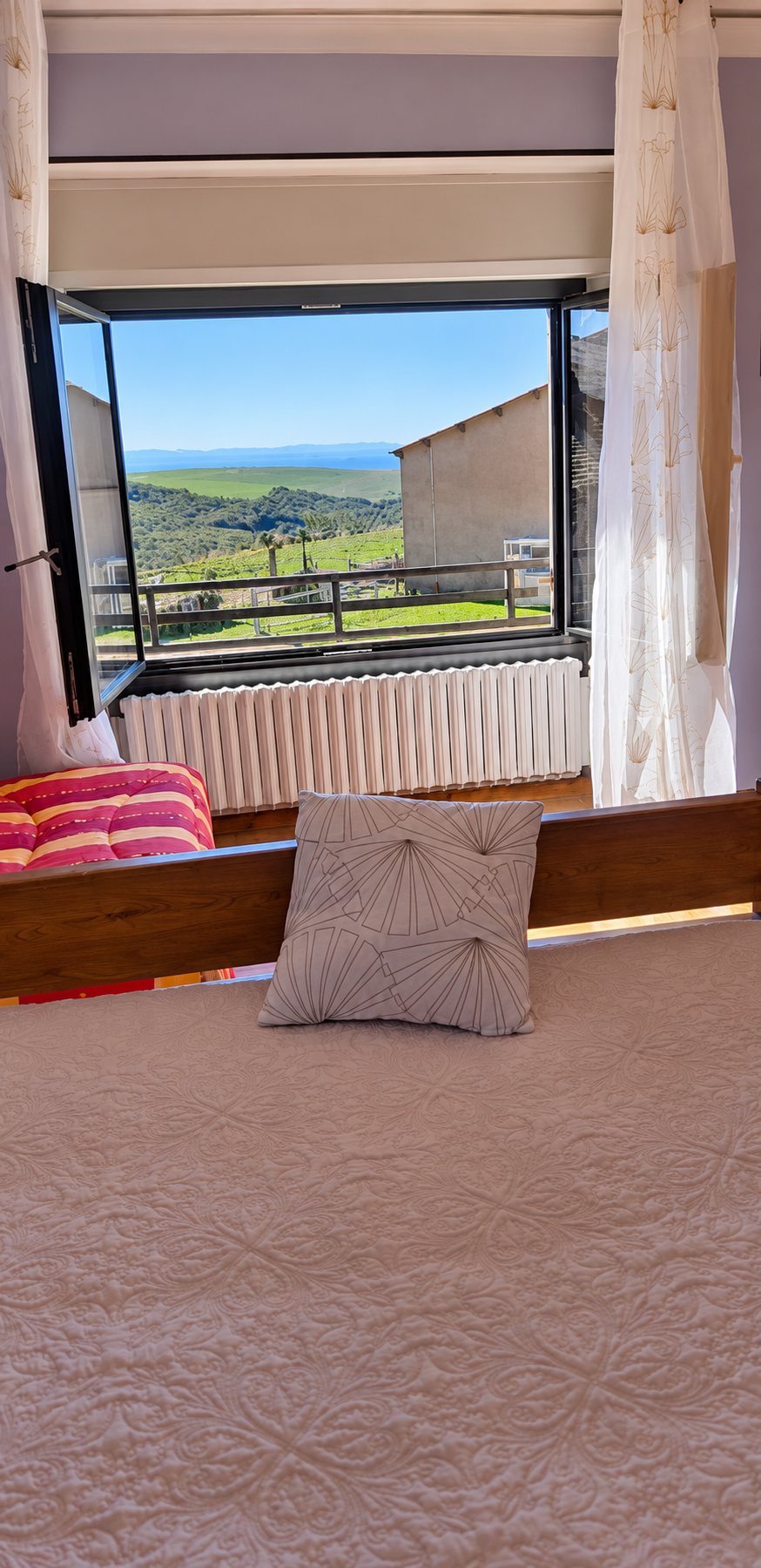 Une chambre avec un lit couvert d'un dessus-de-lit clair et d'un coussin, face à une fenêtre ouverte donnant sur des prés verts et des collines.