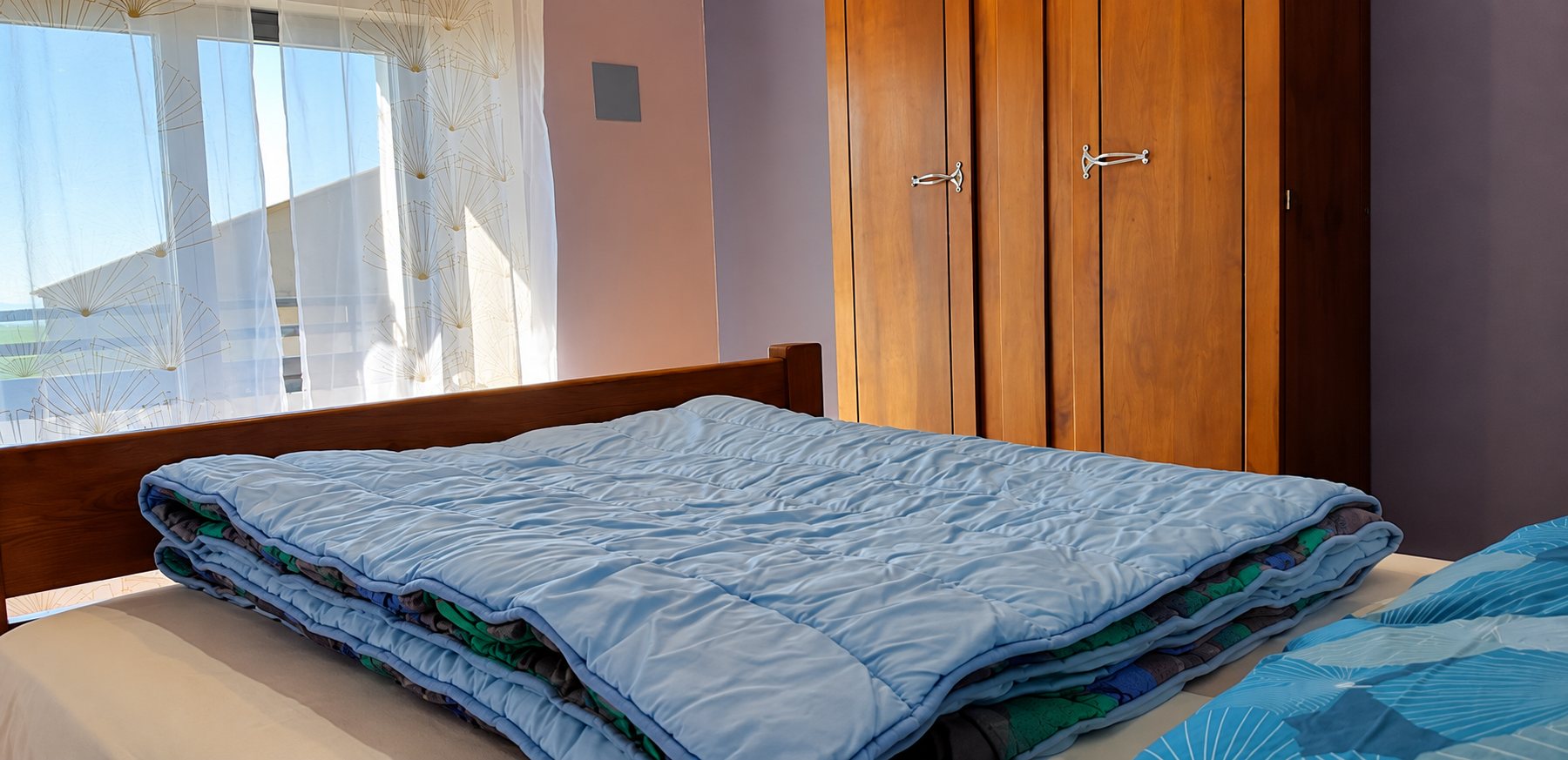 Chambre avec un lit en bois recouvert d'une couette bleue, une grande armoire en bois et une fenêtre aux rideaux blancs en contre-jour.
