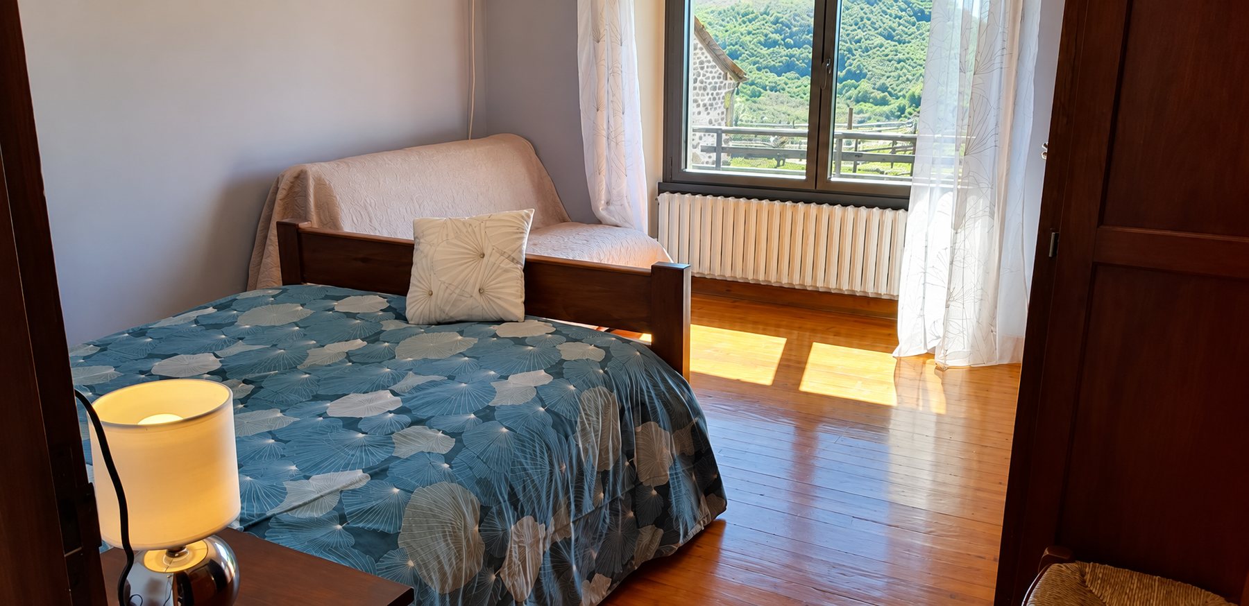 Chambre avec un lit à housse bleue à motifs, une lampe de chevet et un parquet baigné de soleil devant une fenêtre donnant sur une colline verdoyante.