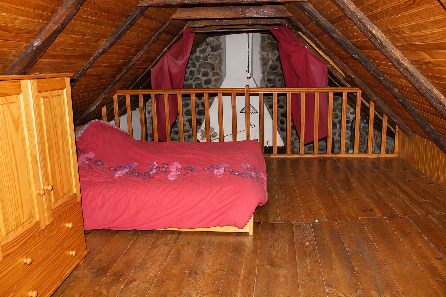 Chambre sous les combles en bois avec un lit recouvert d'une couette rouge, une commode en pin, une balustrade et un mur de pierre au fond.