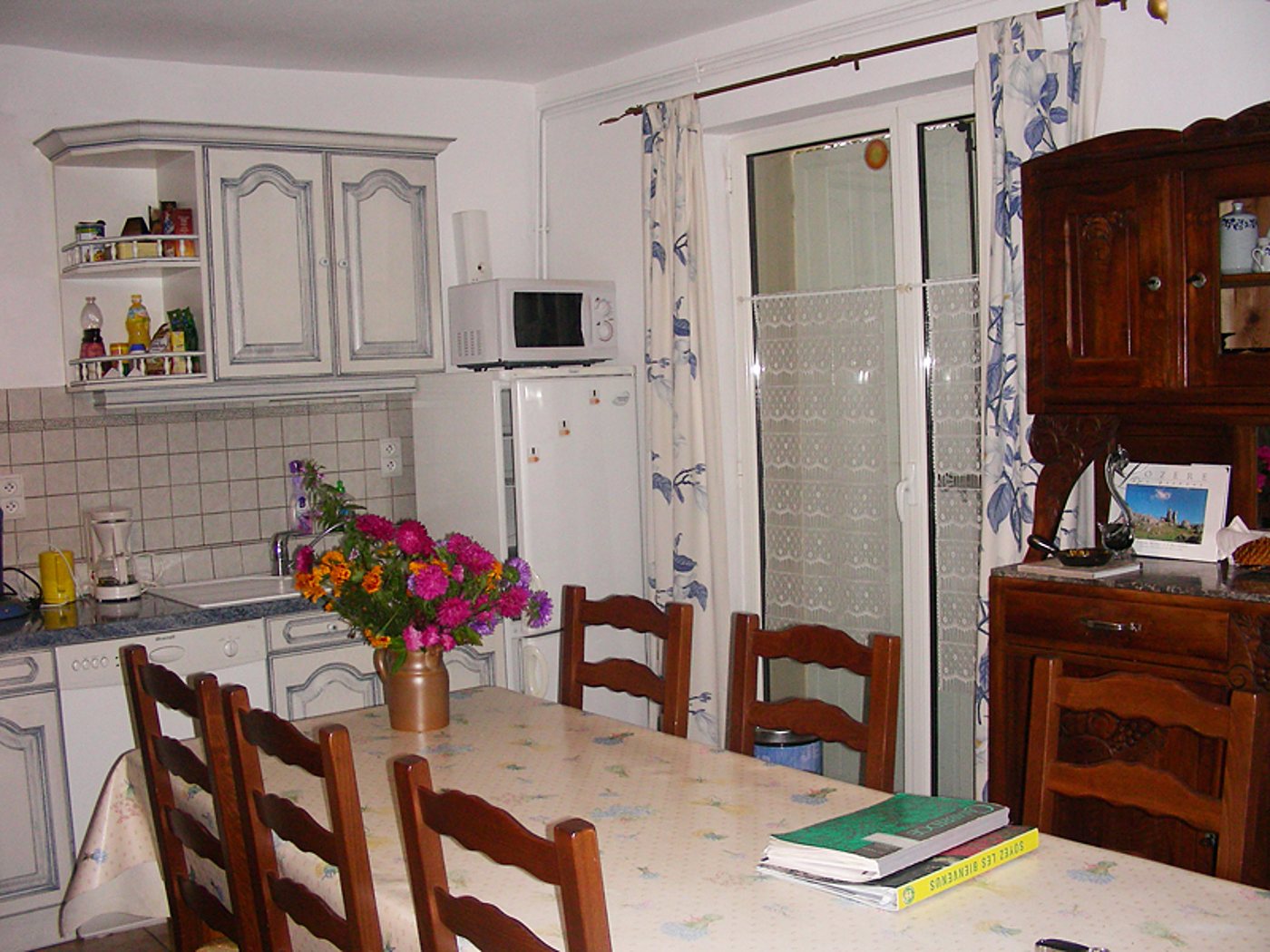Une cuisine familiale avec une table recouverte d'une nappe fleurie, des chaises en bois, un bouquet de fleurs colorées, un buffet ancien et un réfrigérateur près de la porte vitrée.