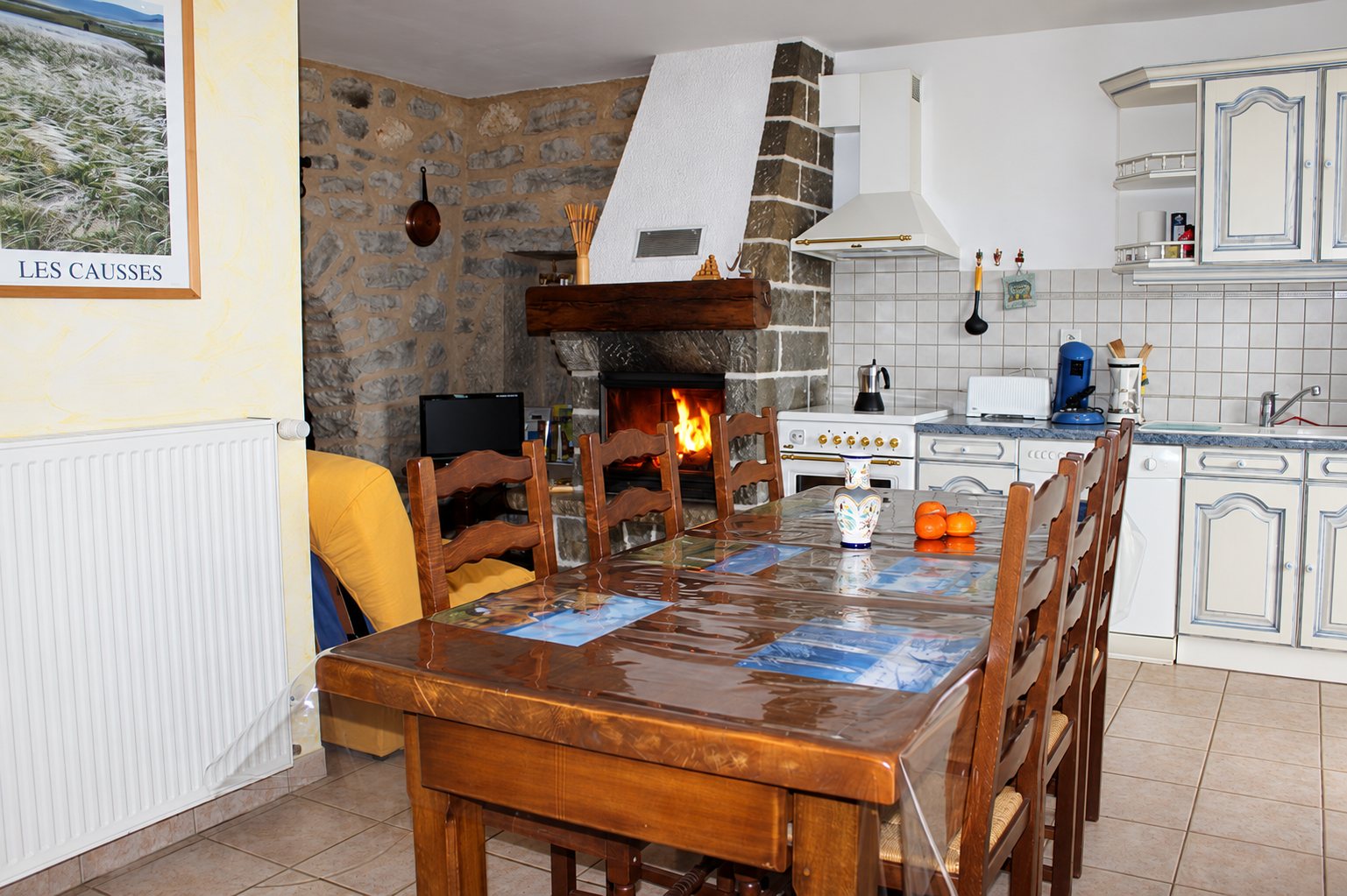 Cuisine rustique d'un gîte avec une grande table en bois et des chaises paillées devant une cheminée en pierre où brûle un feu.