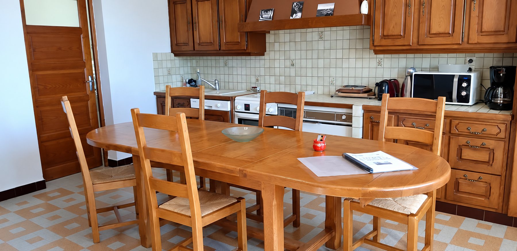 Une cuisine aux placards en bois foncé avec une grande table ovale en bois entourée de chaises à assise en paille, sur un sol carrelé à motifs.