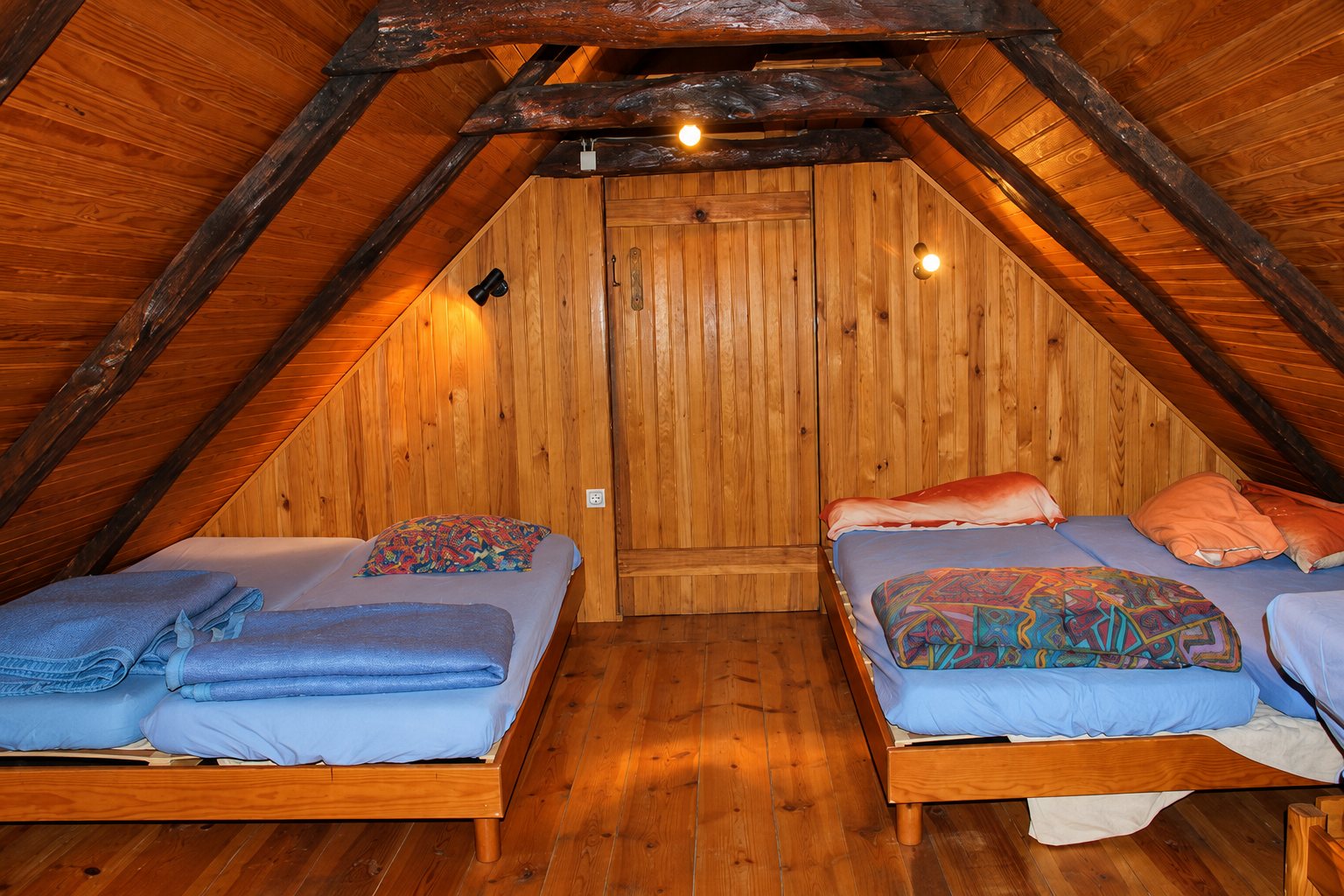 Dortoir sous les combles entièrement habillé de lambris en bois, avec des lits aux draps bleus et de grosses poutres apparentes au plafond.