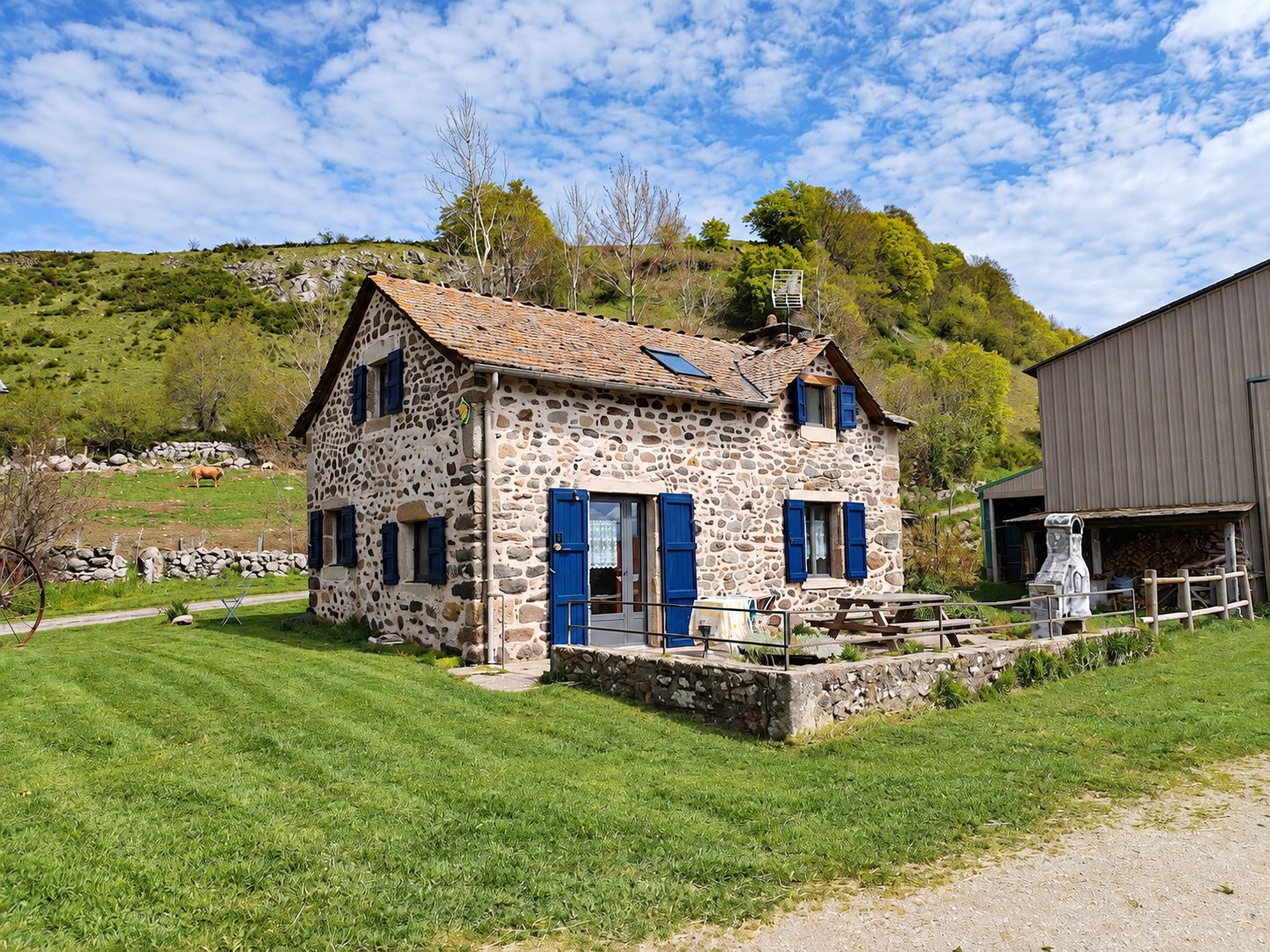 Le Logis des Fées, maison de pierre aux volets bleus et au toit de lauzes, entourée de pelouse sur le plateau de l’Aubrac.