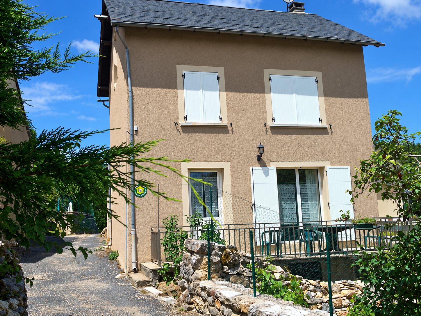 Façade d'une maison en crépi beige à volets blancs, avec une terrasse à garde-corps métallique et un mur en pierre sous un ciel bleu.
