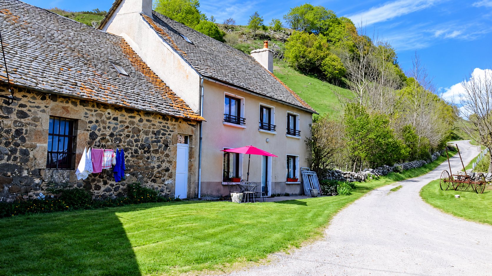 Une maison de campagne en pierre et crépi rose avec du linge qui sèche, un parasol rose et une pelouse verte, au bord d'un chemin dans une vallée.