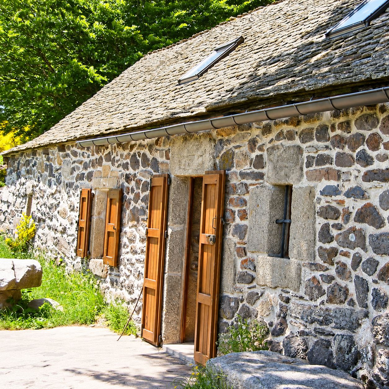 Façade d'une maison en pierres volcaniques au toit de lauzes équipé de fenêtres de toit, avec une porte et des volets en bois ouverts, entourée de verdure.