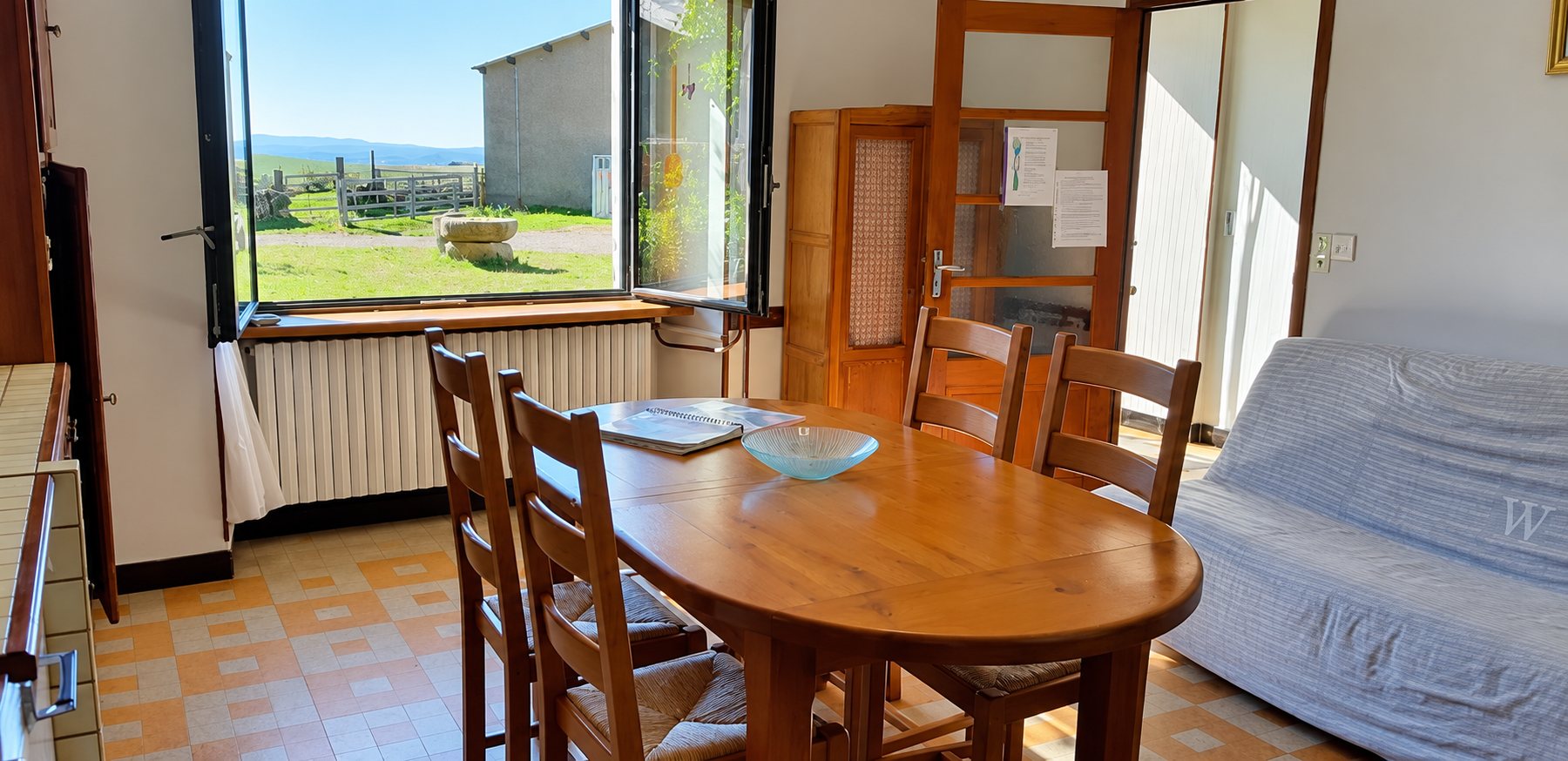 Salle à manger lumineuse avec une table ovale en bois, des chaises, un canapé et une fenêtre ouverte donnant sur la campagne.