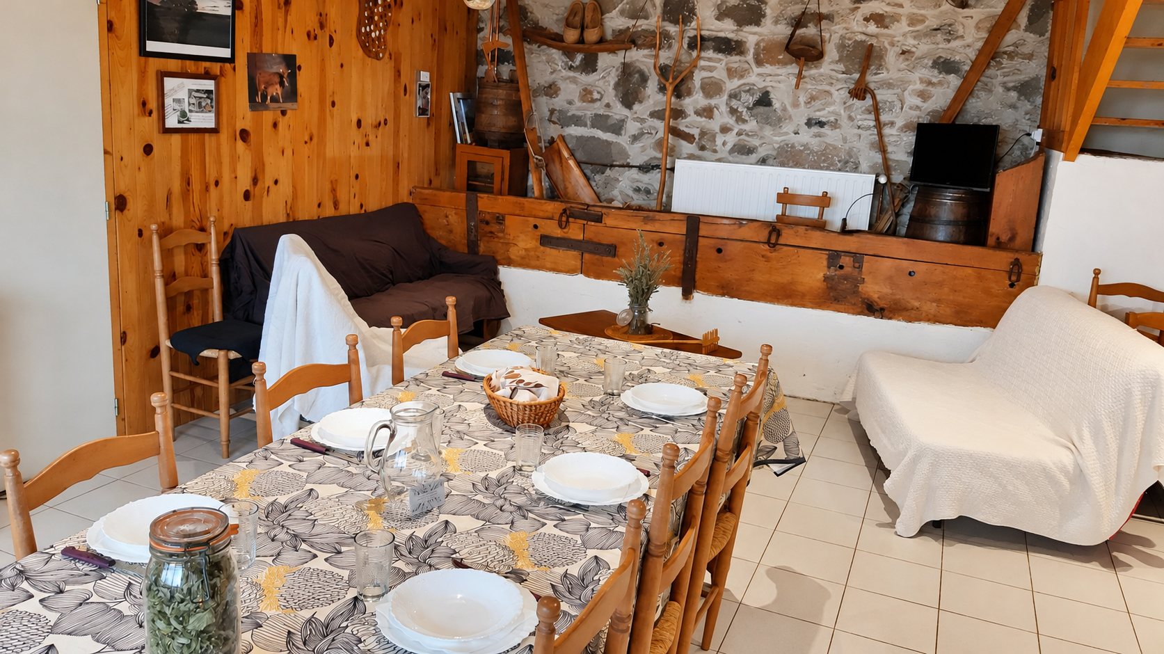 Une salle à manger rustique avec une longue table dressée d'assiettes blanches, des chaises en bois, un canapé et un mur en pierre décoré d'outils anciens.