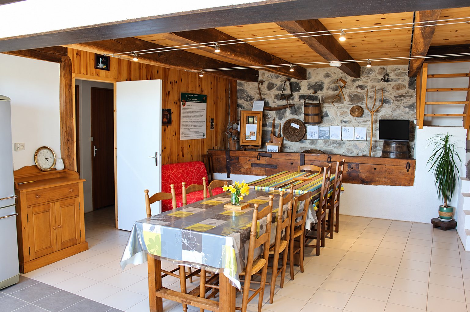 Salle à manger rustique avec une longue table en bois et chaises paillées, poutres apparentes, mur en pierre décoré d'outils anciens et escalier en bois.