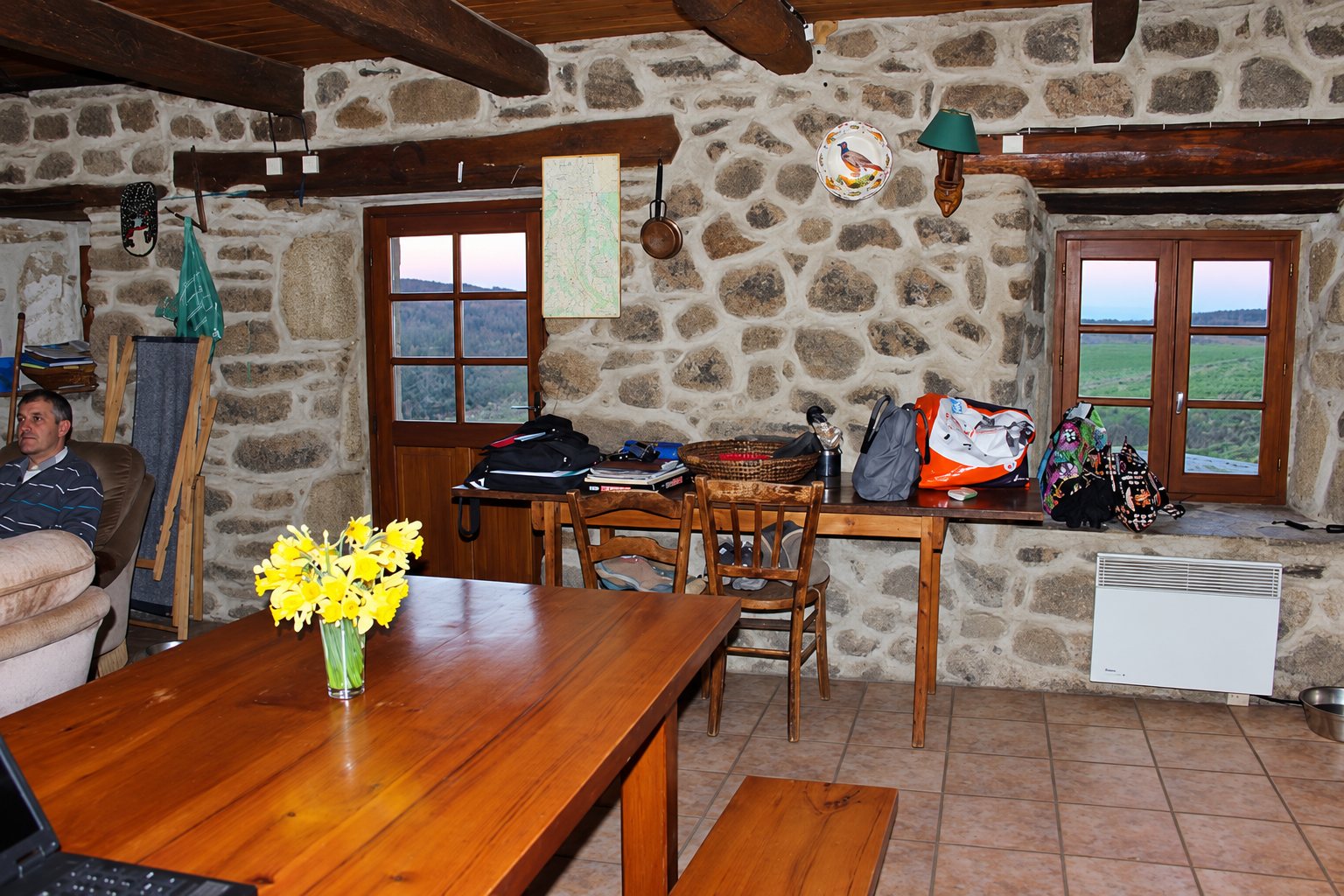 Intérieur d'une salle commune aux murs de pierre apparente, avec une grande table en bois ornée d'un vase de jonquilles, des fenêtres donnant sur la campagne et un homme assis dans un fauteuil à gauche.