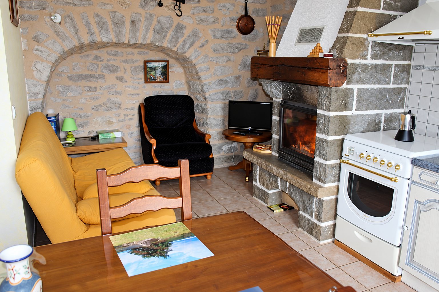 Pièce de vie rustique aux murs de pierre avec cheminée, canapé jaune, fauteuil noir, table en bois et cuisinière blanche.