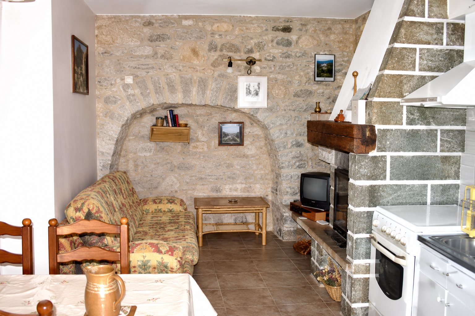 Pièce de vie aux murs de pierre apparente avec un canapé, une table basse, un téléviseur et un escalier en pierre sur la droite.