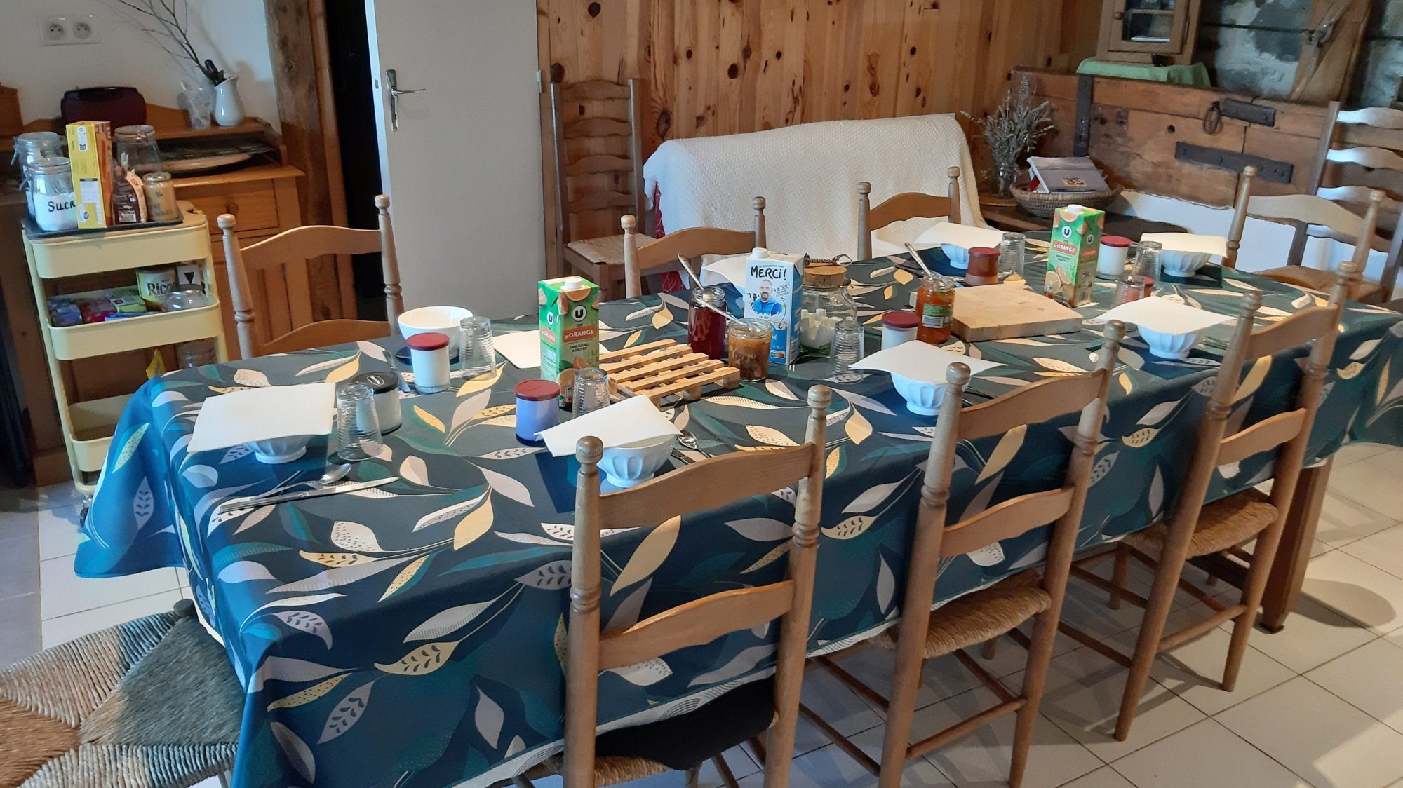 Une grande table recouverte d'une nappe bleue à motifs de feuilles, dressée pour le petit-déjeuner avec bols, confitures et briques de jus, entourée de chaises en bois et paille.