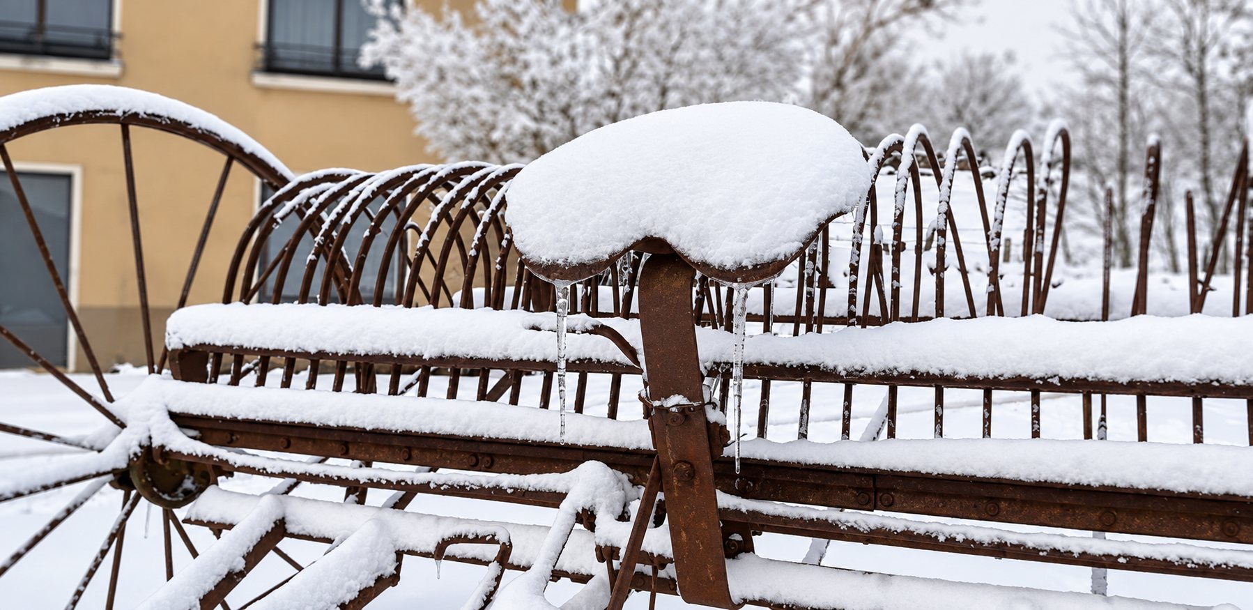 Un ancien râteau-faneur agricole en métal rouillé, dont le siège est couvert de neige et de glaçons, devant un bâtiment en hiver.